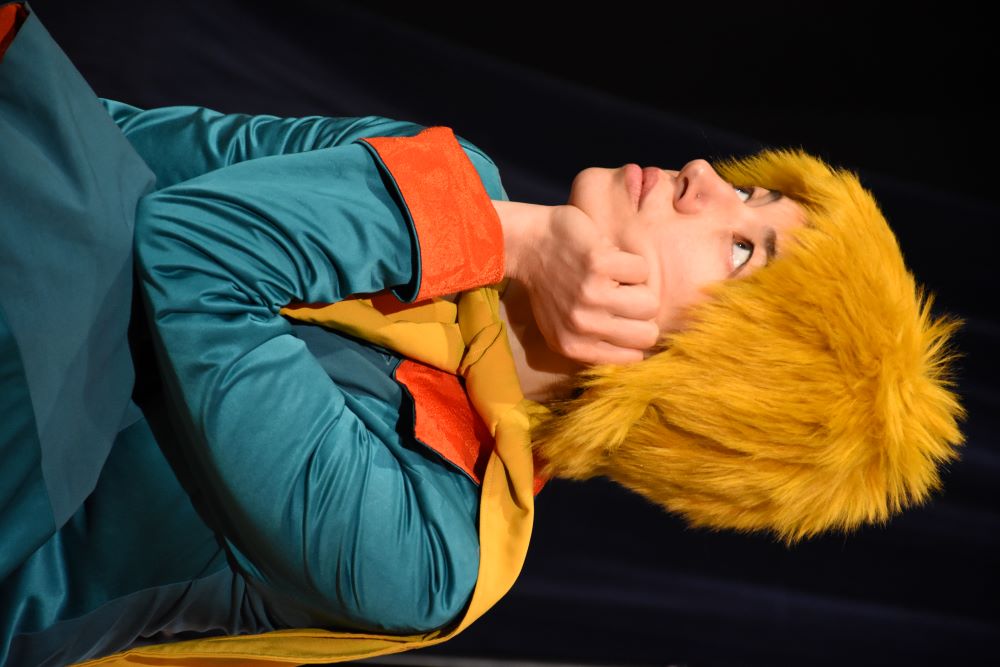 A Golden English Theatre performer as the Little Prince, looking up with a thoughtful smile